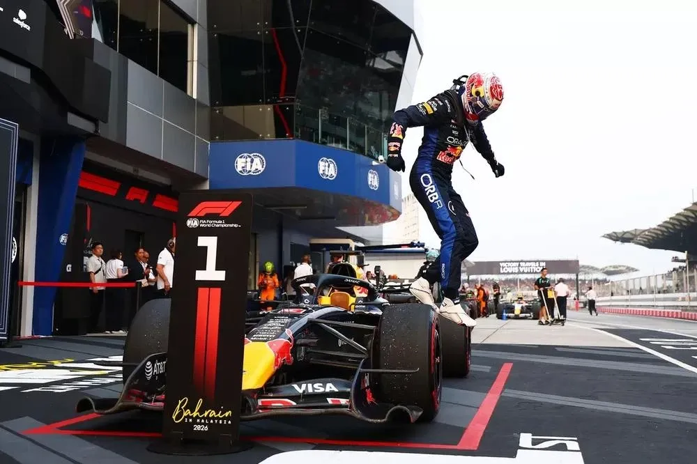 Макс Ферстаппен в гоночном комбинезоне Red Bull стоит на переднем колесе болида после гонки.